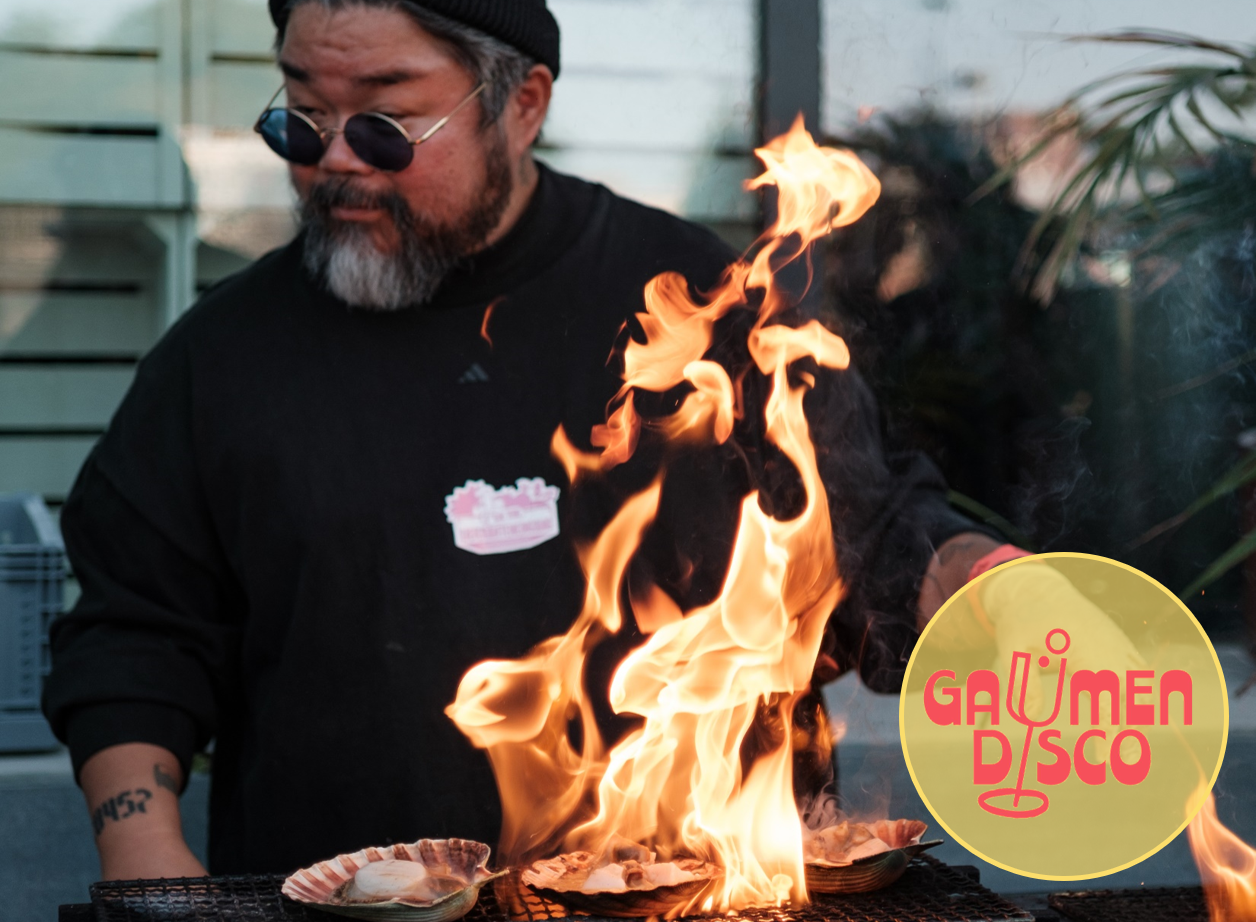 Ein Koch grillt Jakobsmuscheln über offener Flamme bei der Gaumendisco des OPEN MOUTH Hamburg Food Festivals. Flammen lodern spektakulär in die Höhe, rechts unten das Veranstaltungslogo „Gaumendisco“.