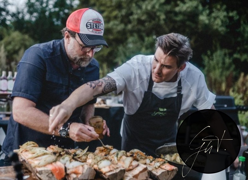 Brian Bojsen and Matthias Gfrörer preparing artfully topped breads outdoors. One wears a chef’s uniform and apron, the other a baseball cap and holds a jar of ingredients. Both work intently side by side at a long wooden table with rustic vegetables in the foreground.