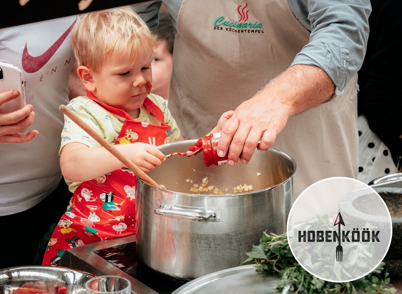 The picture shows a child being lovingly assisted in filling tomato paste; the inscription “Cucinaria” on the adult's apron and the “Hobenköök” logo at the bottom right of the picture are visible.