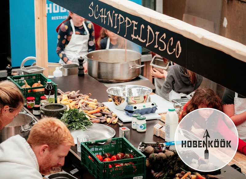 Several people are chopping vegetables together at a long cooking station under a sign reading “Schnippeldisco”. The table is covered with colorful carrots, herbs, beets, and other ingredients. A large cooking pot is visible in the background. The event is hosted by the restaurant Hobenköök.
