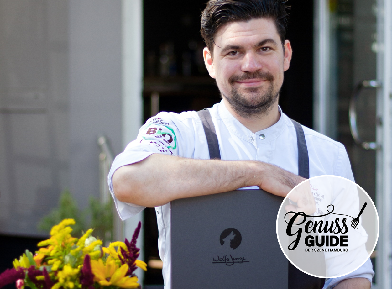 Sebastian Junge, dressed in a white chef's jacket, stands in front of the entrance to his restaurant Wolfsjunge, smiling and leaning on a menu board with the Wolfsjunge logo. A bouquet of flowers is visible on the left; the Genuss Guide Hamburg logo appears in the lower right corner.