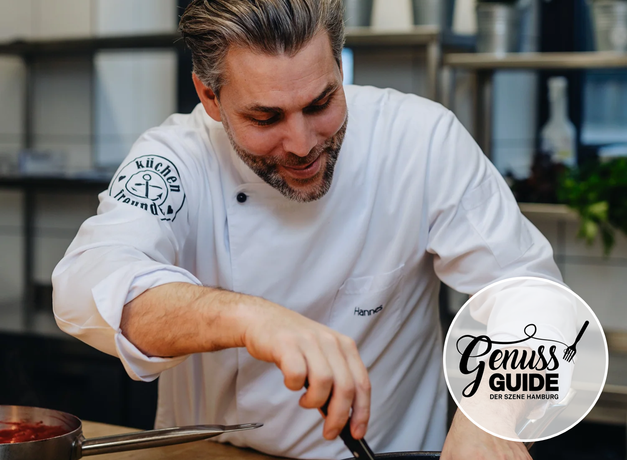 Hannes Schröder is cooking with focus in a professional kitchen. He wears a white chef’s jacket with his name embroidered and the “Küchenfreunde” logo on the sleeve. A saucepan with sauce is visible in the foreground; the Genuss Guide Hamburg logo appears in the lower right corner.