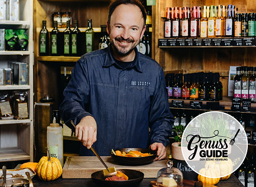Thomas Sampl stands smiling behind a counter, preparing a dish in a pan – surrounded by pumpkins and high-quality food items in his delicatessen shop. Shelves with oils, spices, and spirits are visible in the background. The Genuss Guide Hamburg logo appears in the lower right corner.