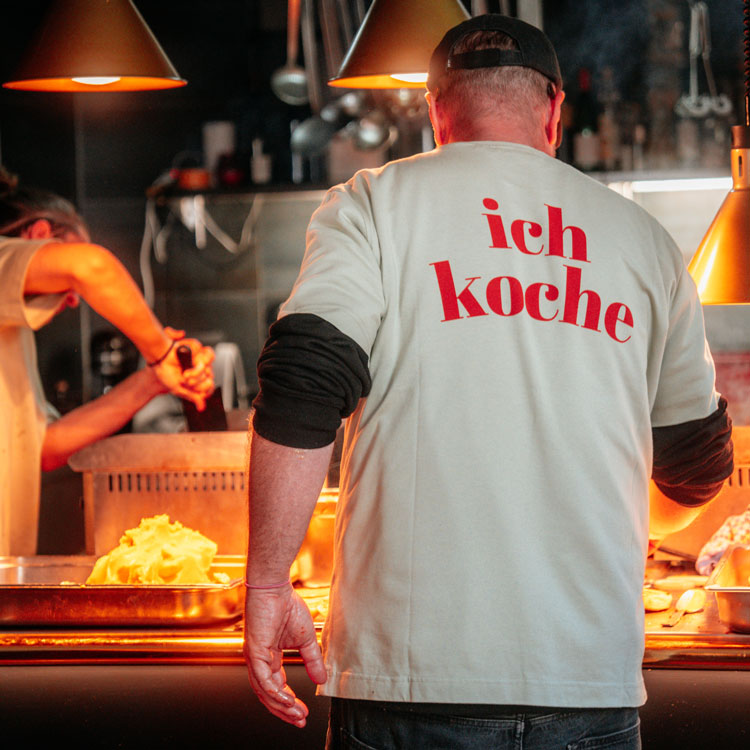 Ein Koch steht am Herd mit dem Rücken zur Kamera. Auf dem T-Shirt prangt der Schriftzug: Ich koche.