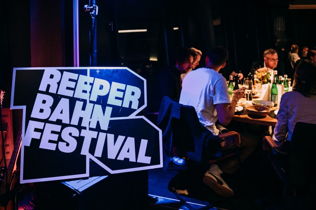 Menschen sitzen an einem Tisch und geniessen Essen und Trinken. Im Vordergrund in das Reeperbahn Festival Logo zu sehen.